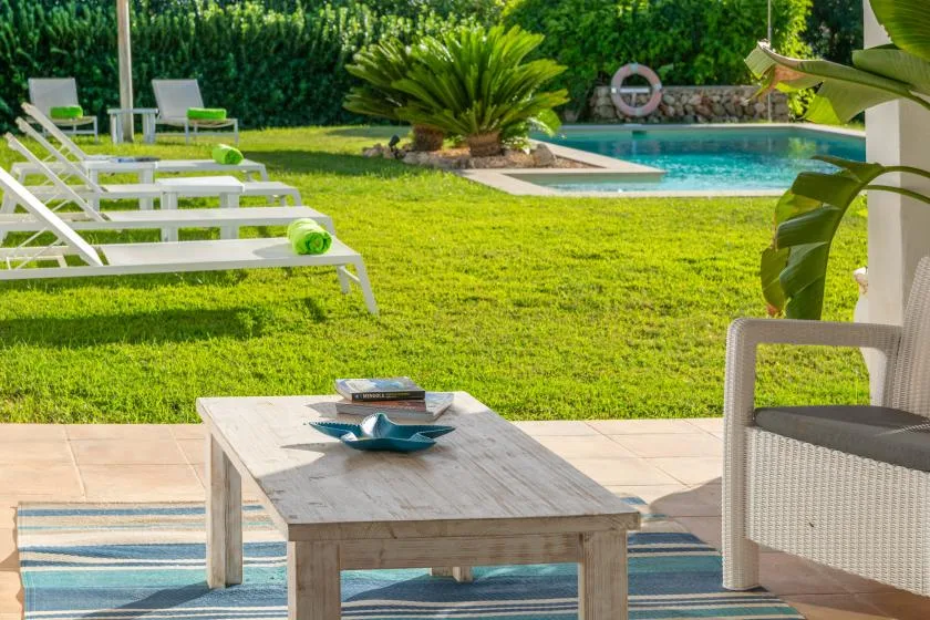 Un patio soleado junto a la piscina tiene asientos, una mesa baja y césped verde.