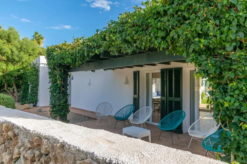 Una zona de estar al aire libre queda bajo una pérgola cubierta de enredaderas junto a un edificio blanco.