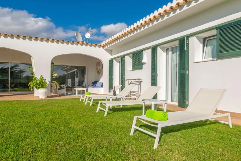 Un patio soleado junto a una casa blanca, con asientos sobre el césped y contraventanas verdes.