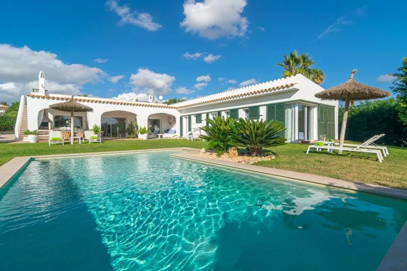 Una piscina está frente a una casa blanca con patio y vegetación.