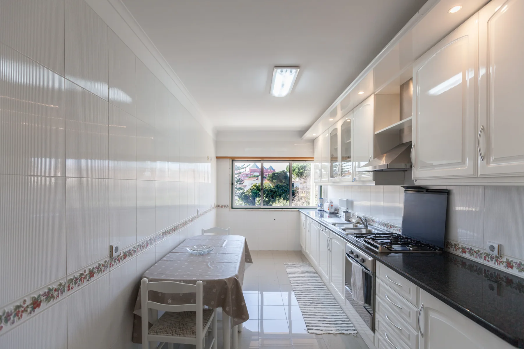 Una cocina alargada con azulejos, armarios blancos, encimeras oscuras y una mesa pequeña.