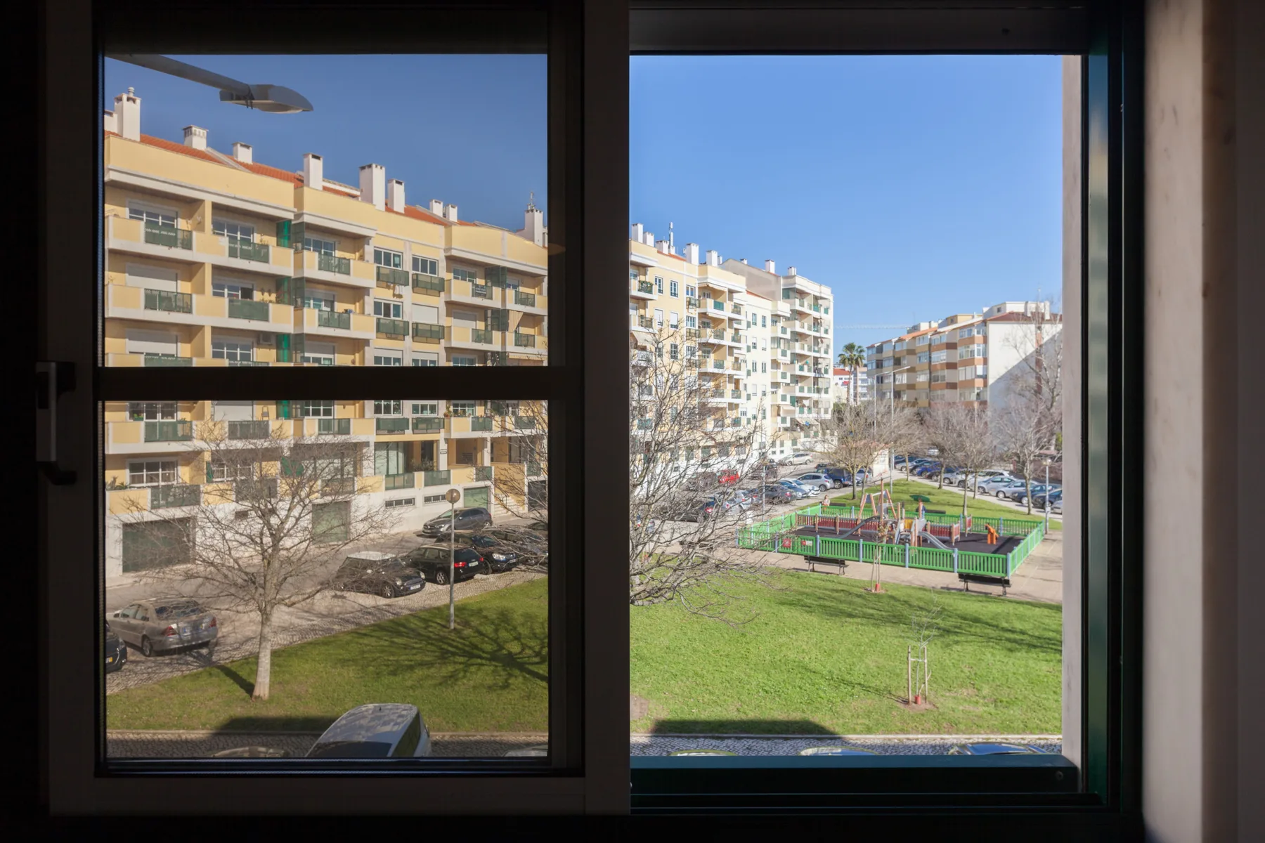 Vista de edificios de apartamentos, un patio verde y una pequeña cancha deportiva a través de una ventana.