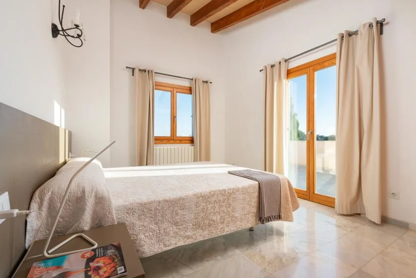 Un dormitorio luminoso con una cama junto a ventanas altas y cortinas beige.