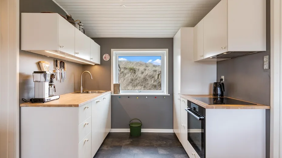 Una cocina pequeña con gabinetes blancos, encimeras y una ventana con vista a dunas cubiertas de hierba.