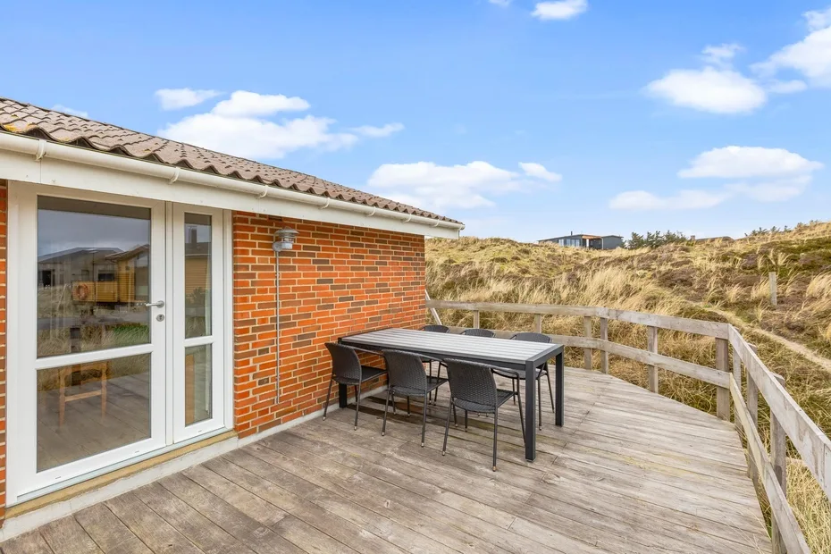 Una terraza exterior junto a una pared de ladrillo tiene una mesa y asientos frente a dunas cubiertas de hierba.