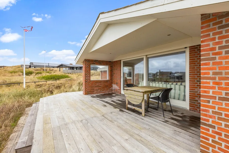Una terraza de madera junto a una casa de ladrillo tiene una mesa y sillas bajo un techo.