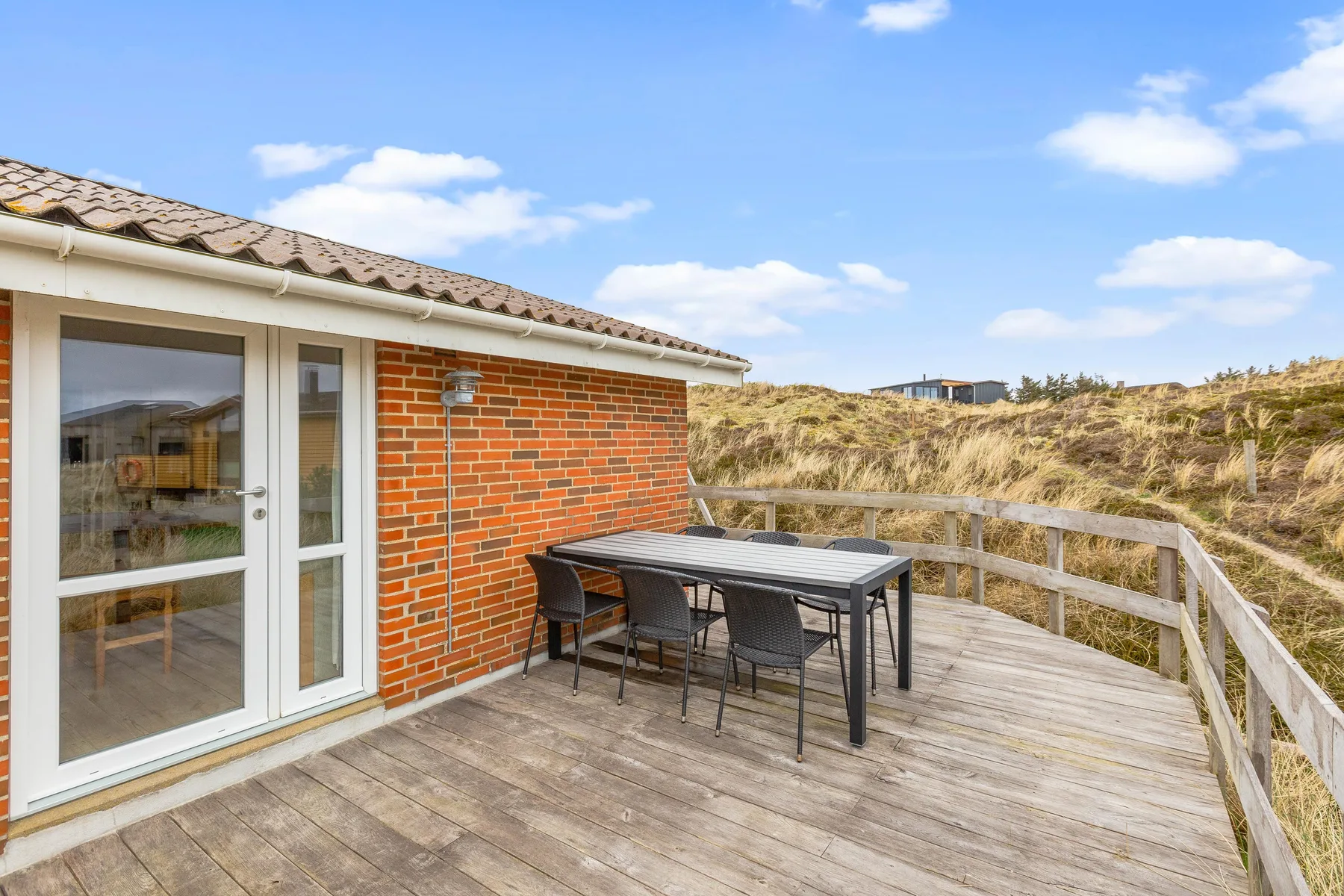 Una terraza de madera junto a la casa tiene asientos al aire libre y vistas a dunas cubiertas de hierba.