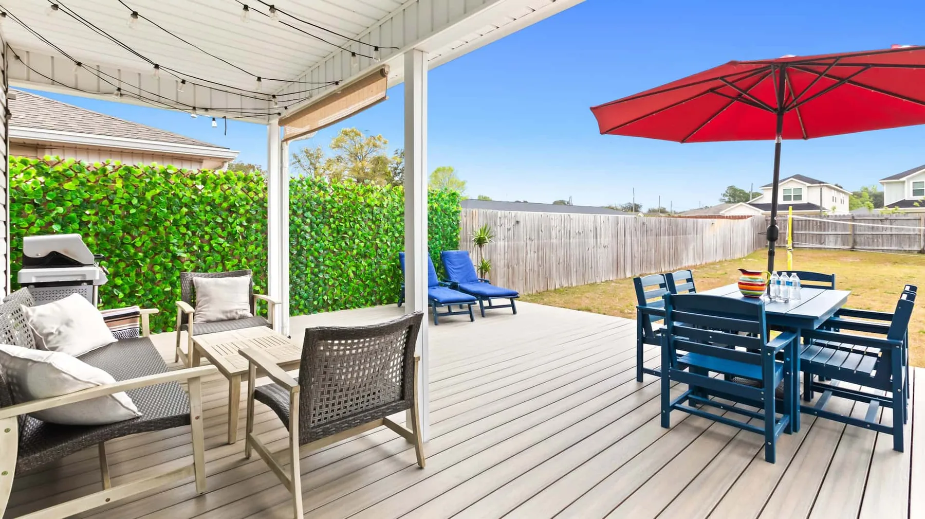 A covered outdoor deck has seating beside a dining table under a red umbrella.