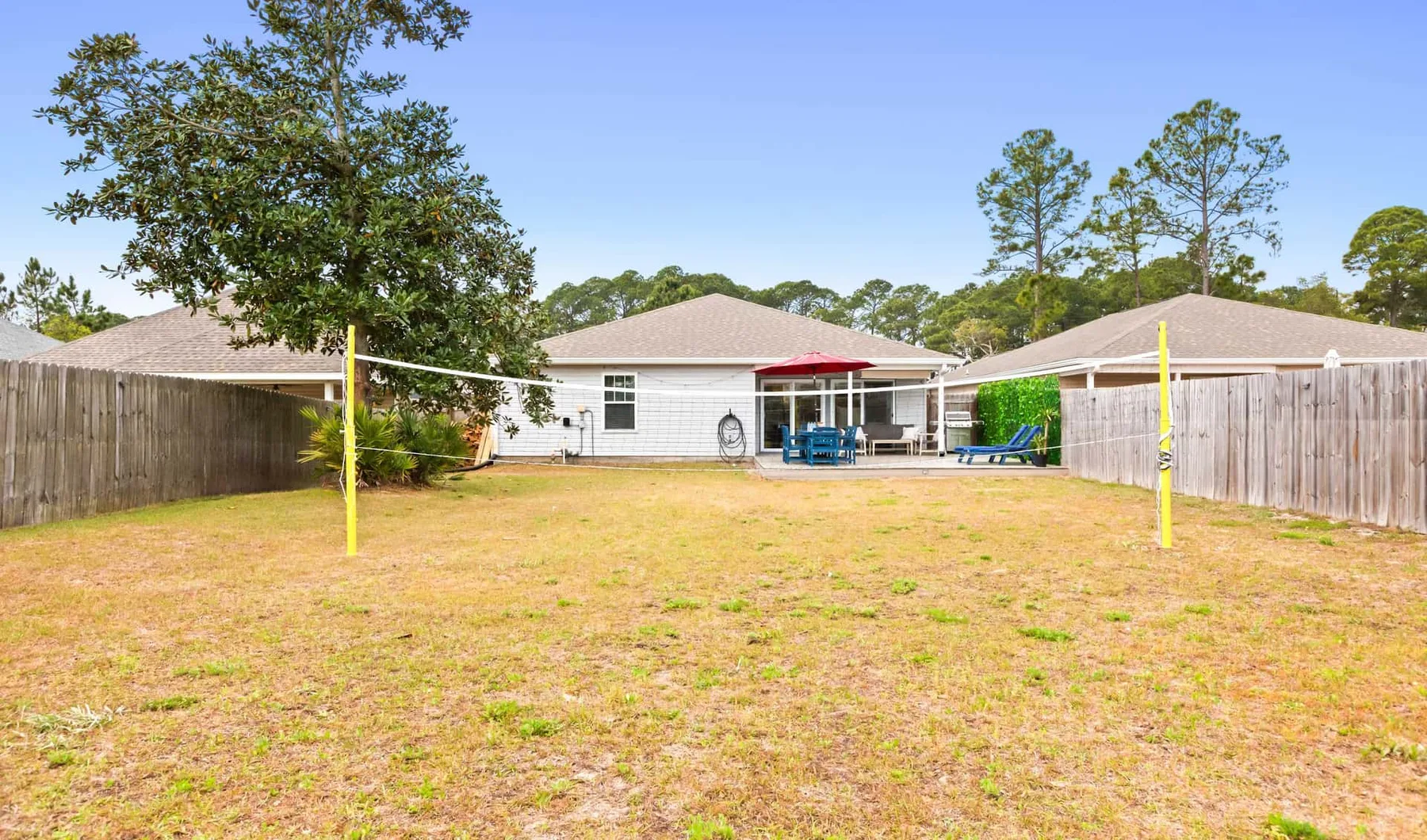 A fenced backyard with a lawn, a volleyball net, and a patio behind the house.