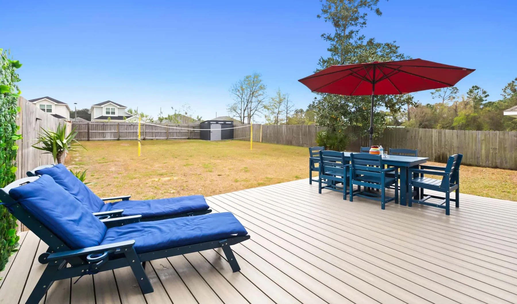 A backyard deck has blue lounge seating and a dining table beneath a red umbrella.