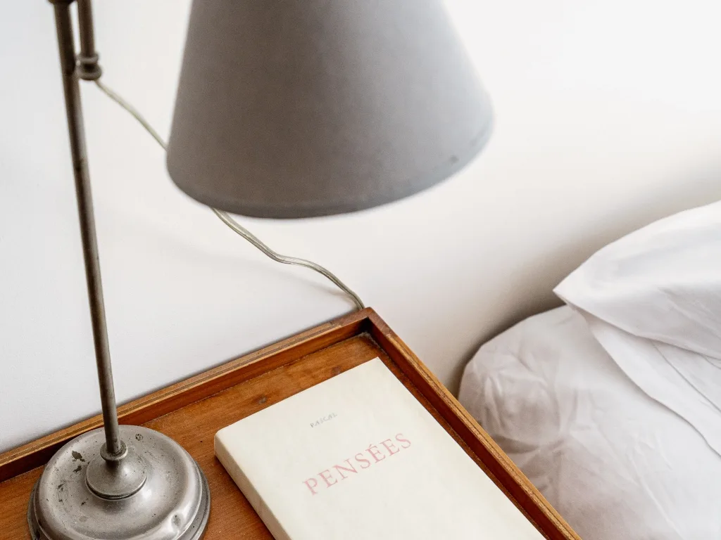 Una mesita de noche tiene un libro junto a una cama y una lámpara metálica.