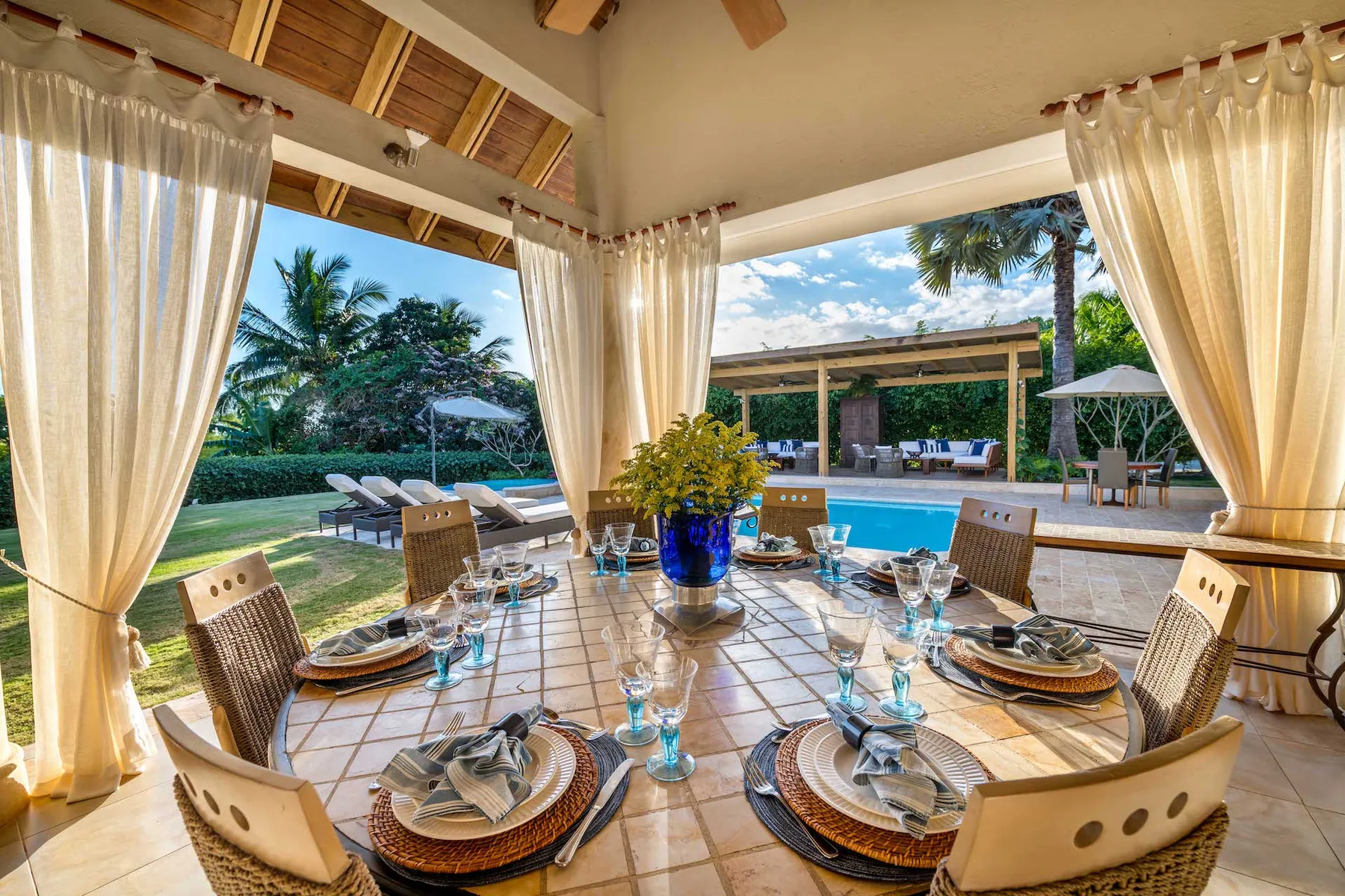 Un comedor cubierto se abre a un jardín junto a la piscina, con la mesa preparada para comer.