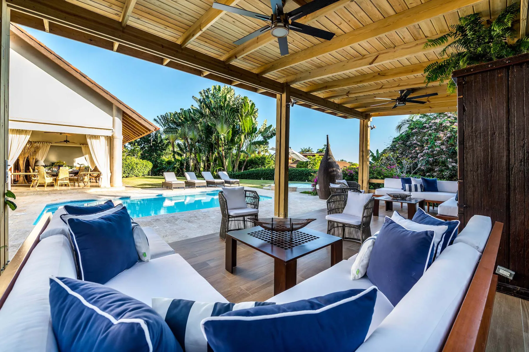 Una sala exterior cubierta da a una piscina, con asientos acolchados y una mesa baja.