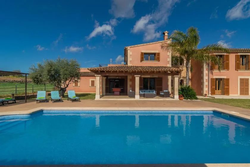 Una piscina frente a una casa, con asientos y palmeras.
