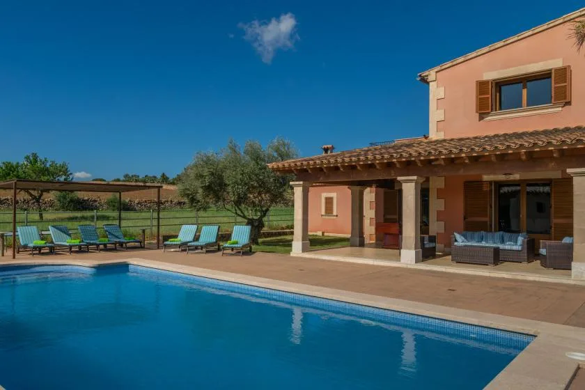 Una piscina exterior está junto a una casa con un patio cubierto y tumbonas.