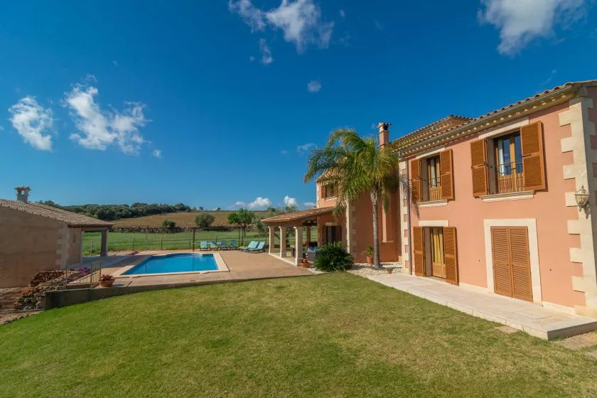 Una casa y una piscina bordean un césped bajo un cielo azul despejado.