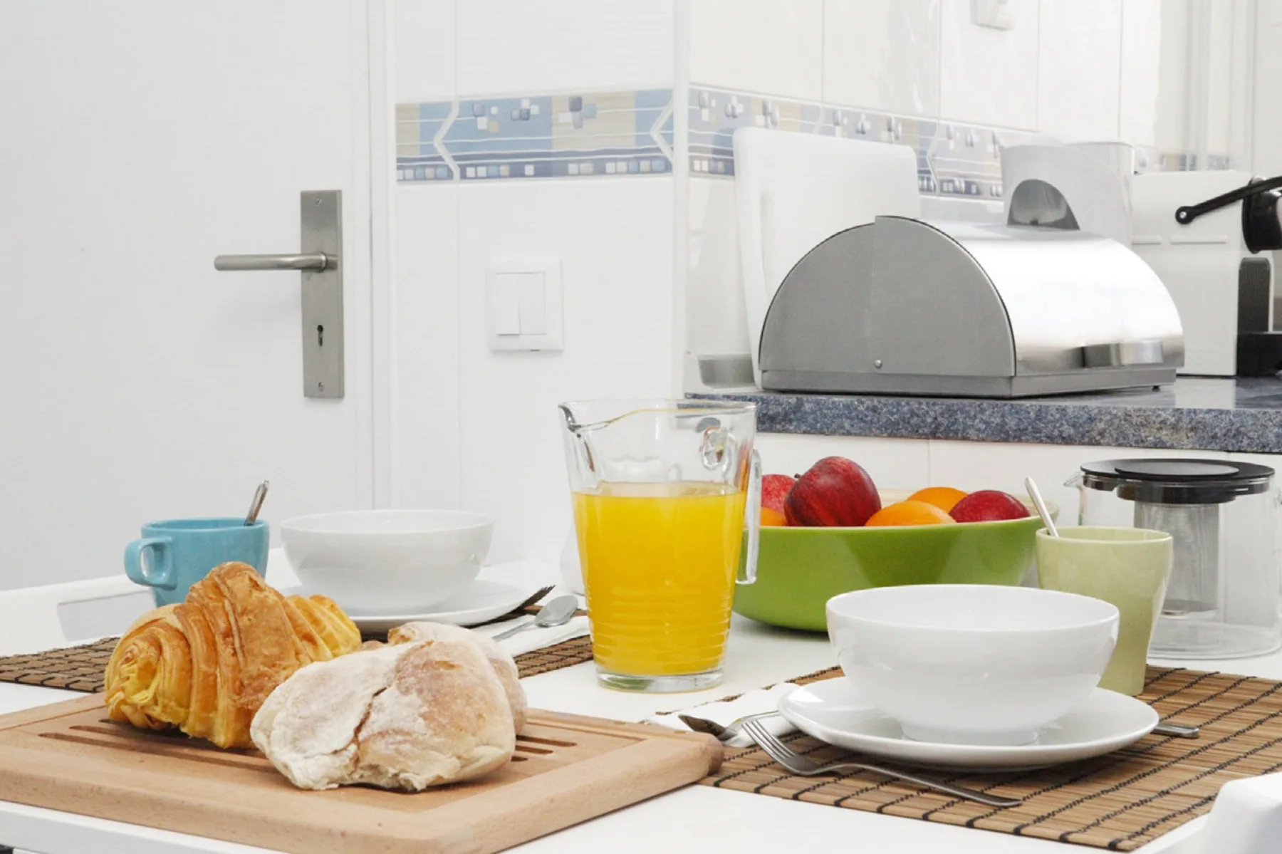 A bright kitchen counter holds breakfast food, juice, and a bowl of fruit.