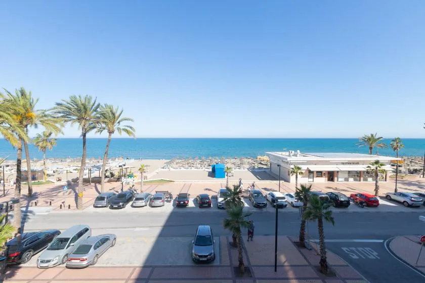 A coastal street and parking area overlook a sandy beach and the sea.