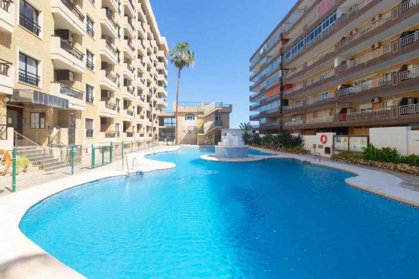 An outdoor swimming pool sits between apartment buildings under a clear blue sky.