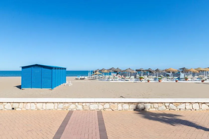 A sandy beach with a blue hut and rows of sun loungers beneath parasols.