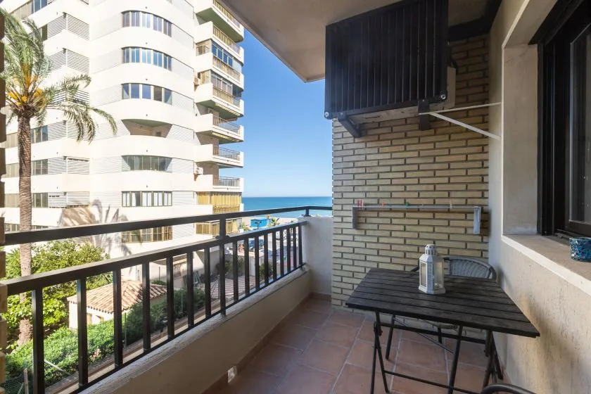 A balcony with a table and seating overlooks nearby buildings and the sea.