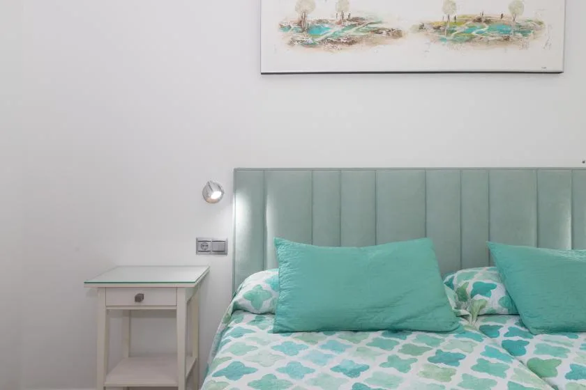 A bedroom with a bed dressed in turquoise and white bedding beside a small table.