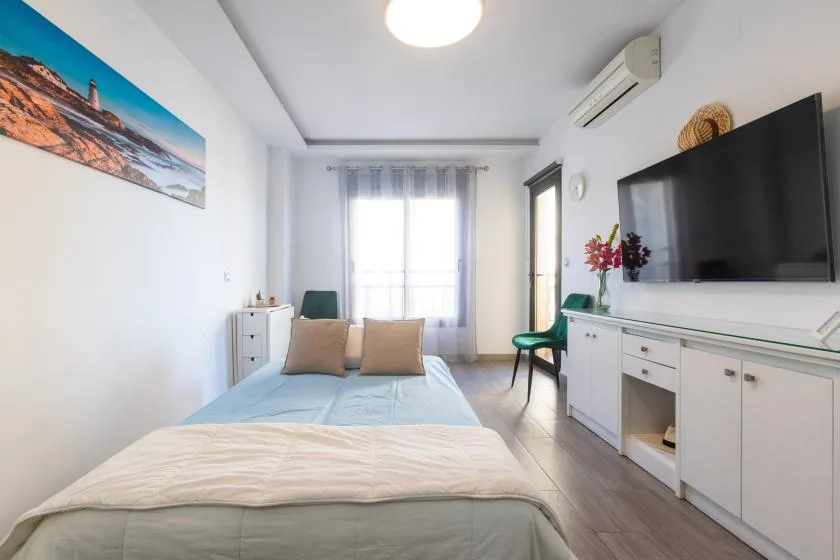 A bright bedroom with a bed, a window, and a television mounted above storage cabinets.