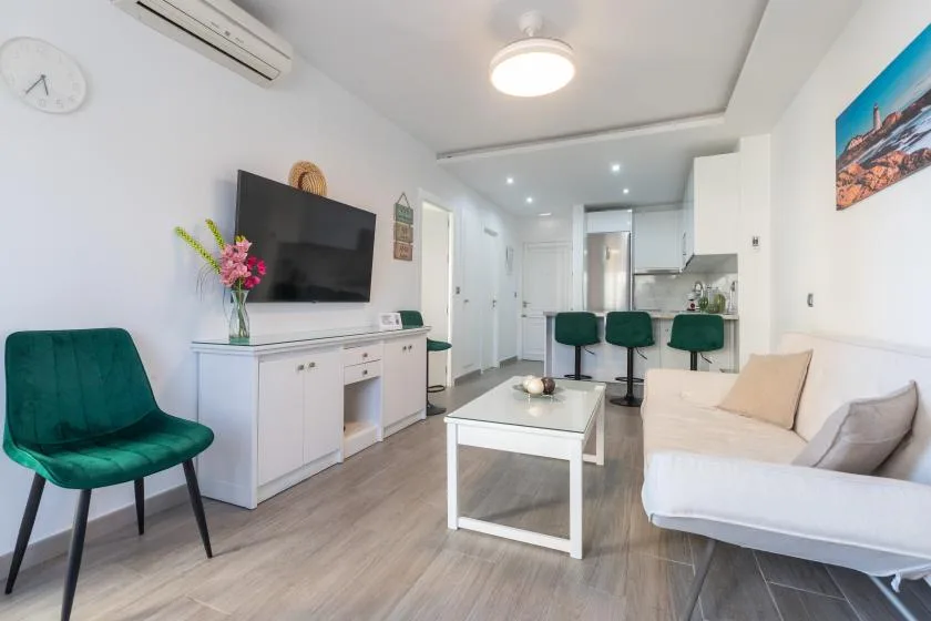 A bright living room with a television, coffee table, and seating beside a kitchen.