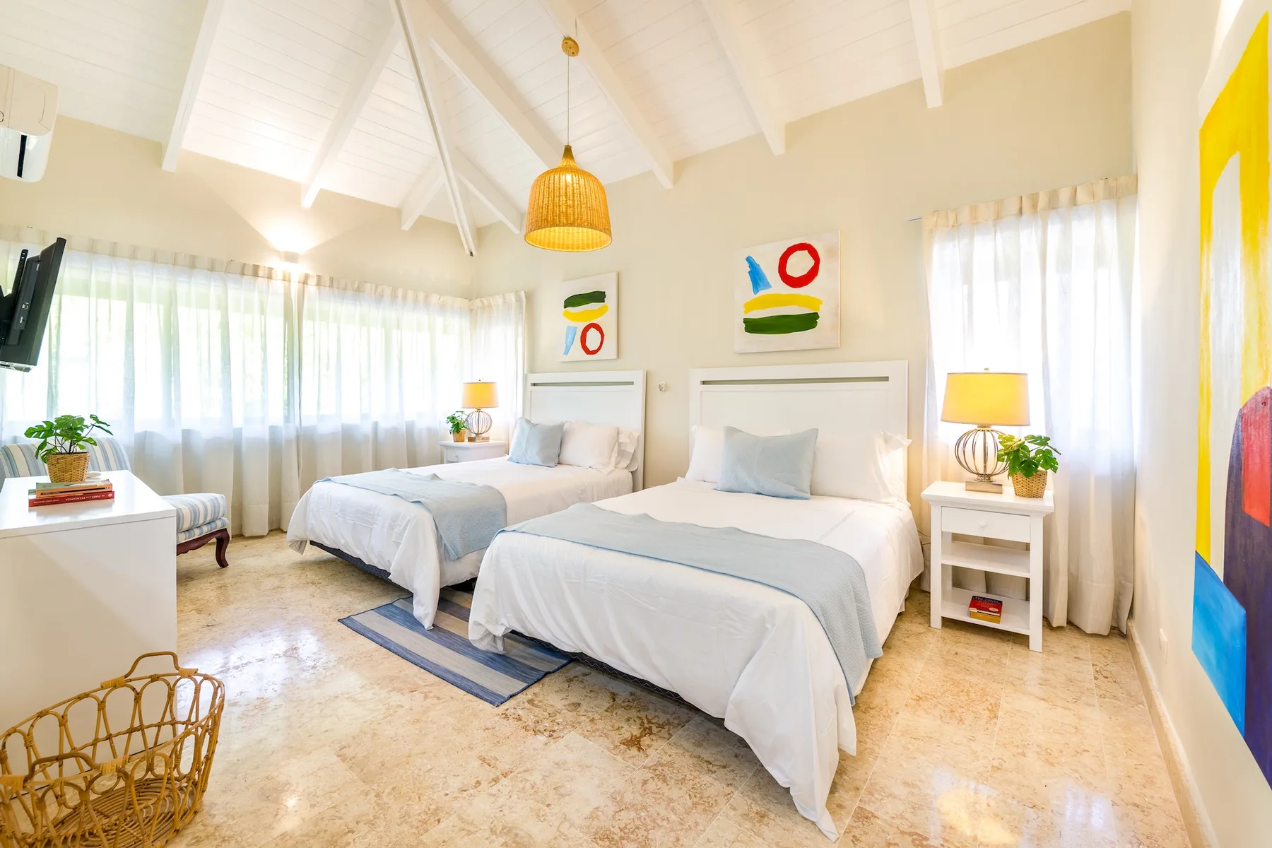 Un dormitorio luminoso con dos camas, ropa de cama clara y grandes ventanas con cortinas.
