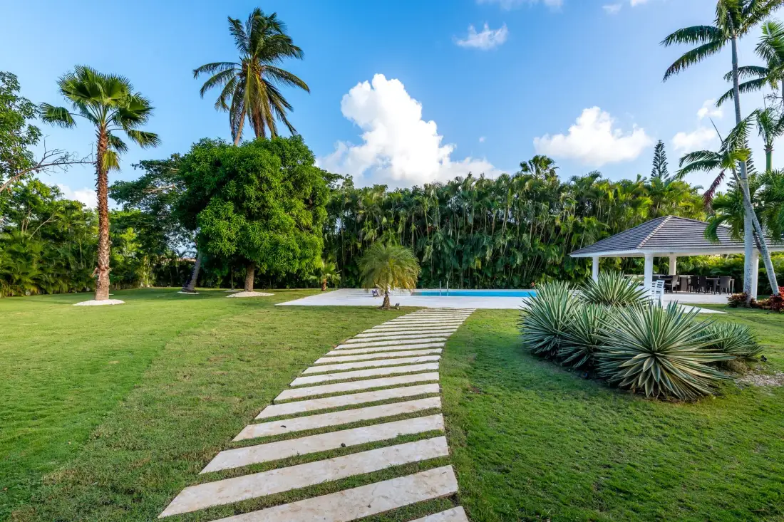 Un sendero de jardín cruza el césped hacia una piscina y un pabellón abierto.