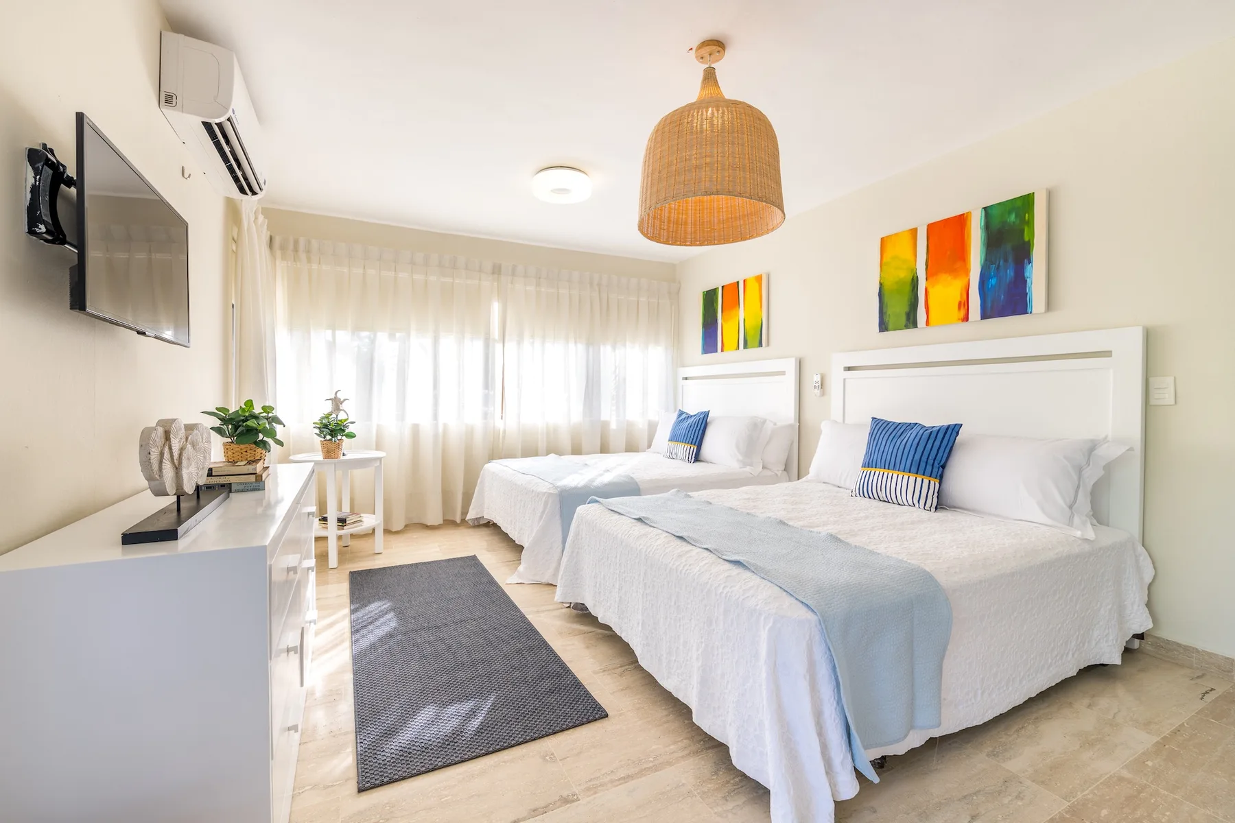 Un dormitorio luminoso con dos camas, cortinas translúcidas y arte colorido en la pared.