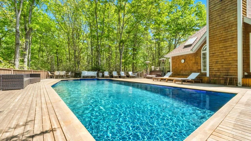 Una piscina al aire libre junto a una terraza de madera, rodeada de árboles.