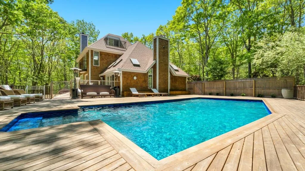Una piscina está junto a una terraza de madera, con una casa y árboles al fondo.