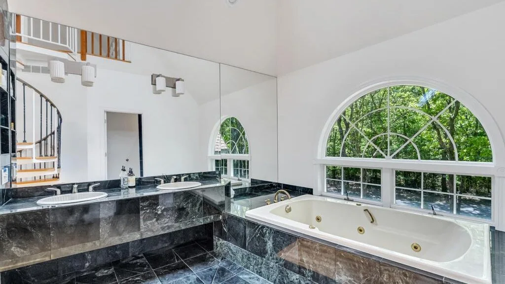 Un baño luminoso con una bañera grande bajo una ventana arqueada.