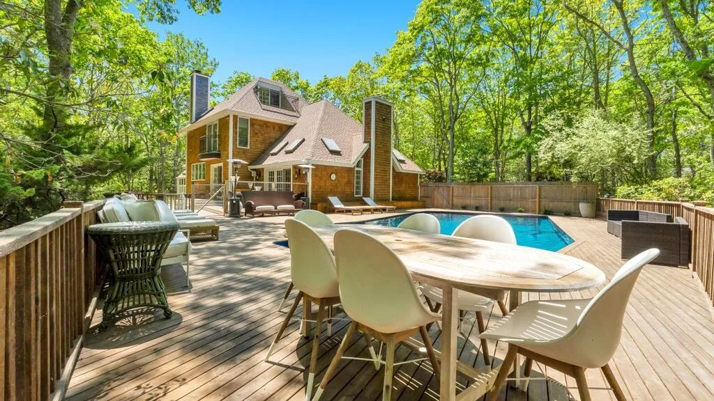 Una terraza de madera con muebles de comedor al aire libre da a una piscina junto a una casa entre árboles.