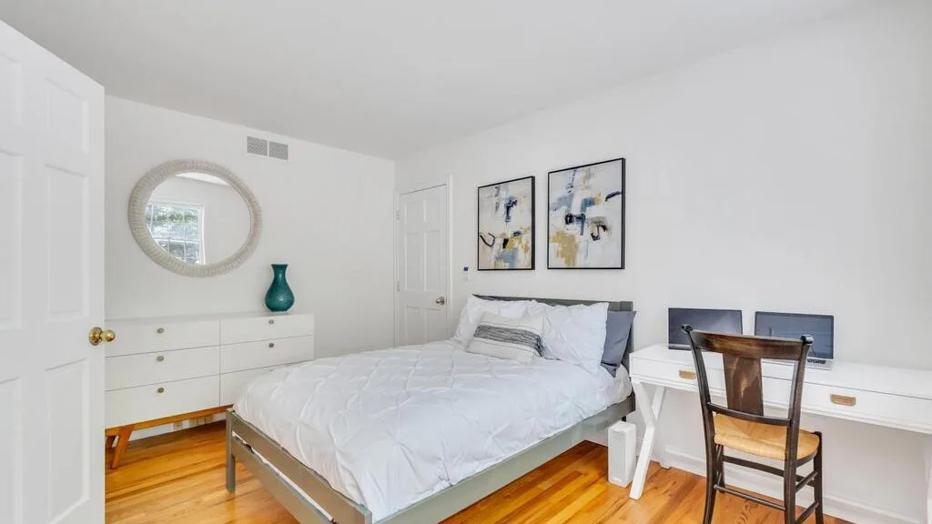 Un dormitorio luminoso con cama, cómoda y escritorio bajo cuadros enmarcados.