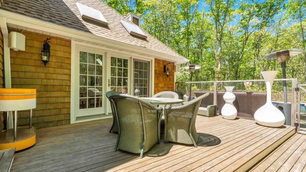 Una terraza de madera junto a una casa tiene asientos al aire libre y una mesa.