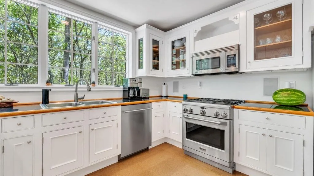 Una cocina luminosa con gabinetes blancos, ventanas grandes y electrodomésticos de acero inoxidable.