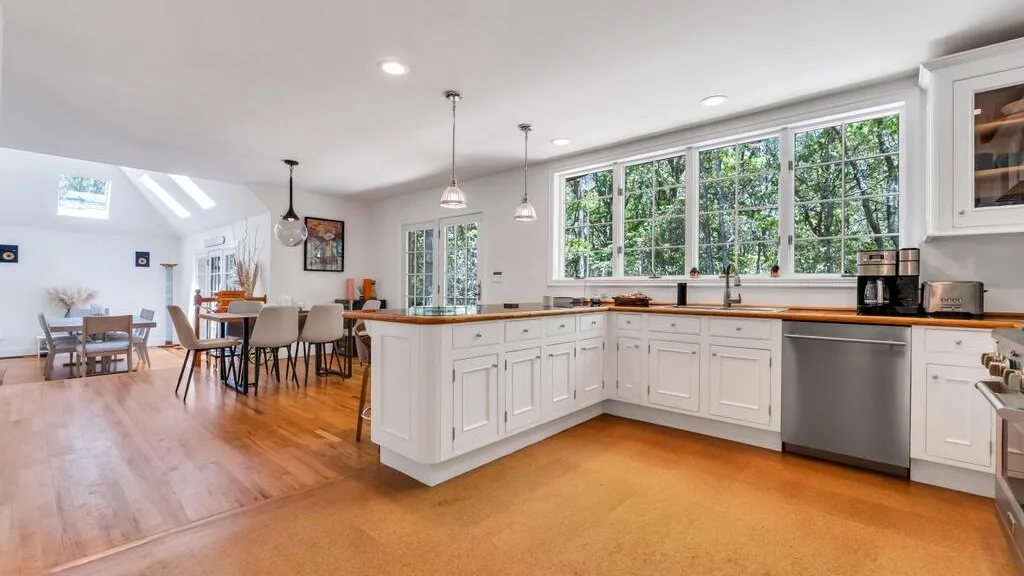 Una cocina luminosa con gabinetes blancos, encimeras de madera y ventanas grandes.