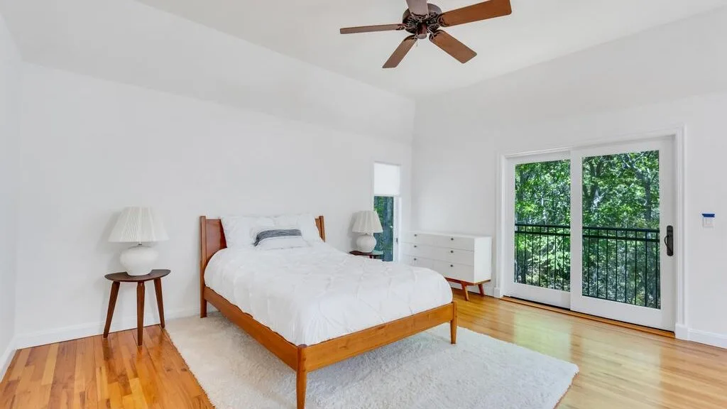 Un dormitorio luminoso con cama de madera, ventilador de techo y puertas de vidrio hacia la vegetación.