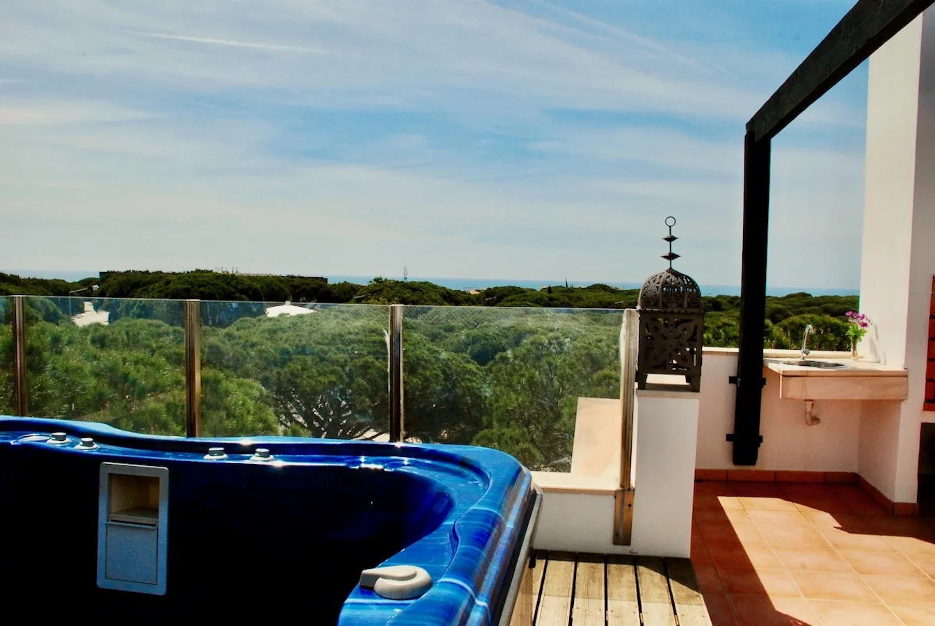 A rooftop terrace with a blue hot tub and glass railing overlooks greenery.