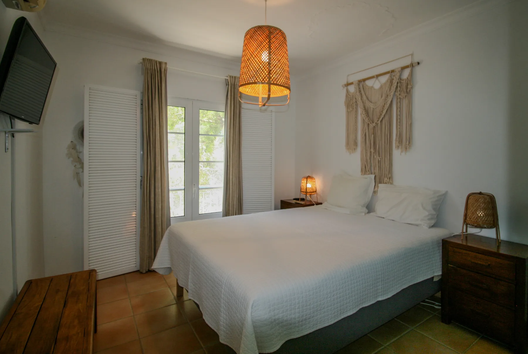 Un dormitorio con una cama bien hecha, ventanas con persianas y una cálida lámpara colgante.