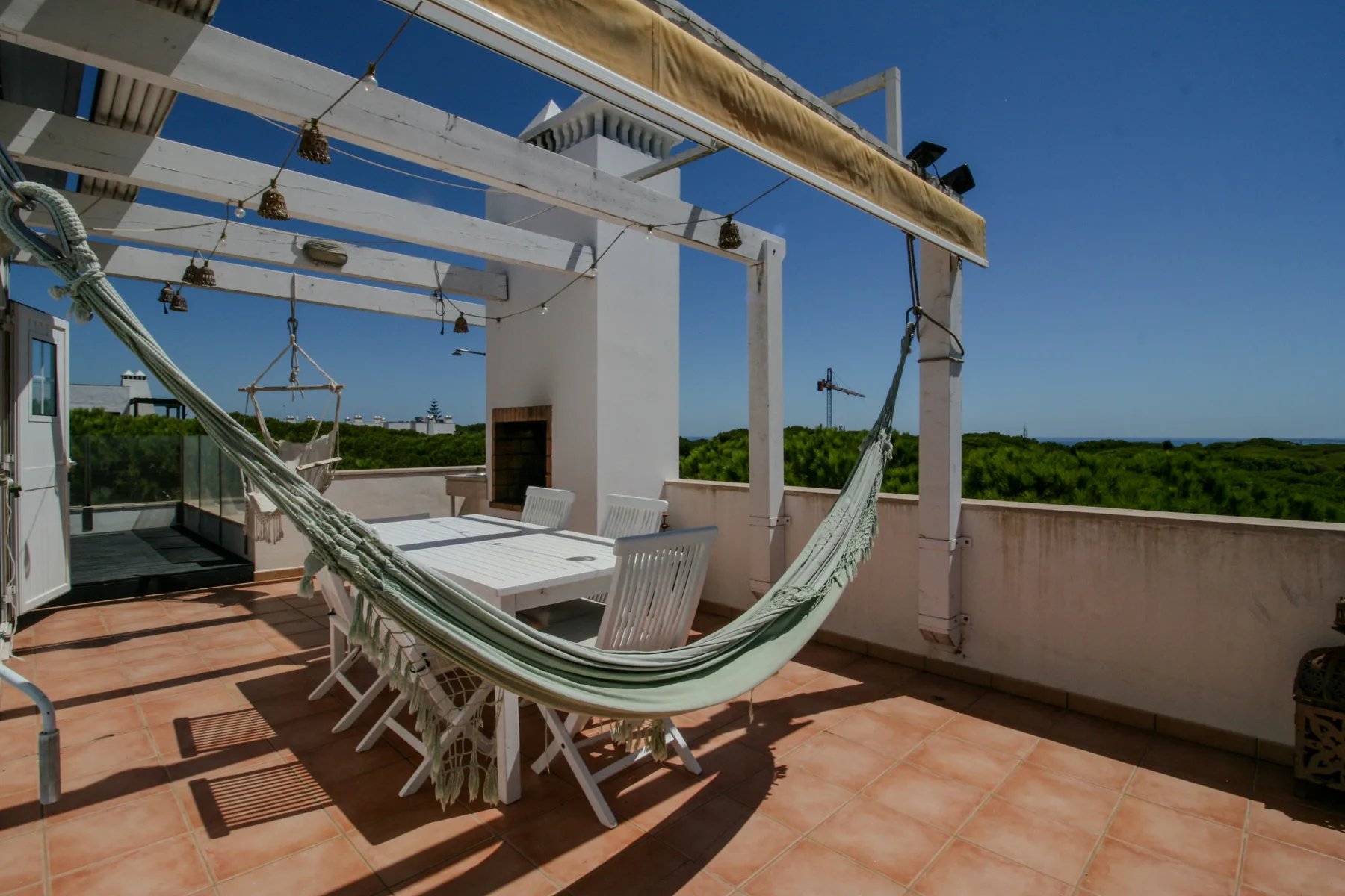 A rooftop terrace has a dining table and a hammock beneath a pergola.