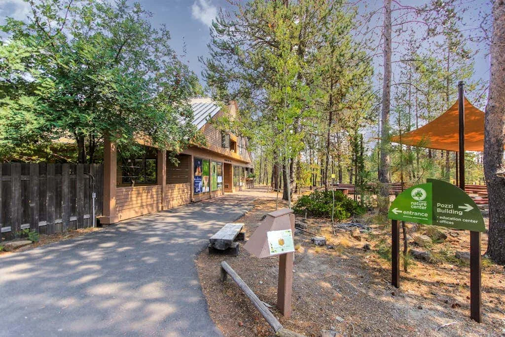 Entrada exterior arbolada con un sendero pavimentado, un edificio de madera y señales.