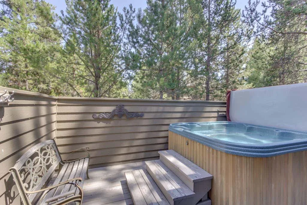 Una bañera de hidromasaje al aire libre está junto a una terraza de madera y asientos, rodeada de árboles.