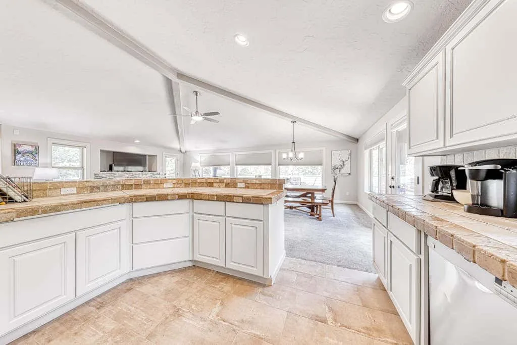 Una cocina luminosa con gabinetes blancos, encimeras de piedra y un comedor.