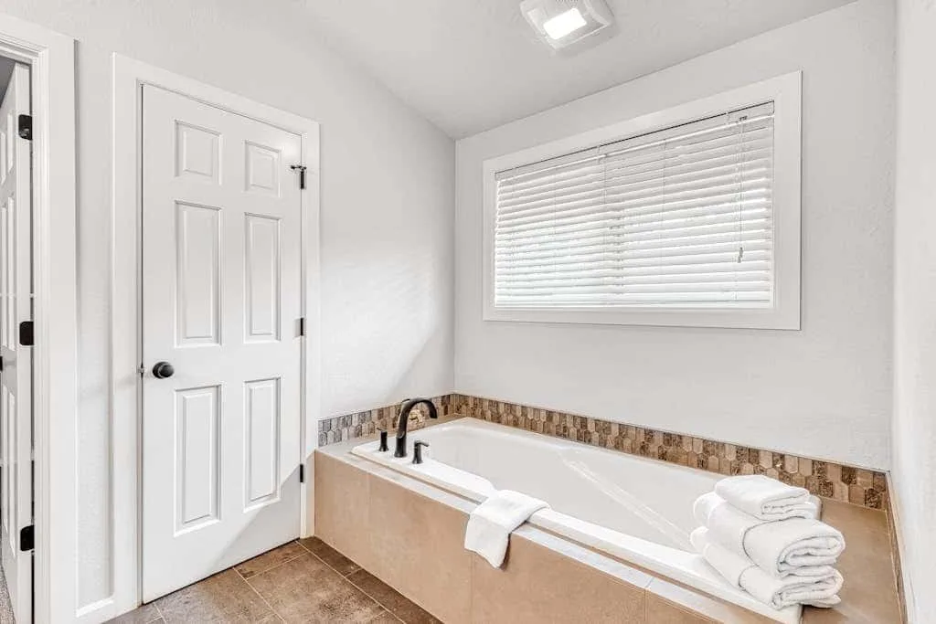 Un baño luminoso con una bañera bajo una ventana con persianas cerradas.