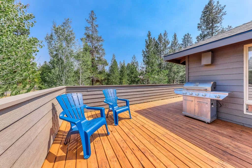 Una terraza de madera tiene asientos azules y una parrilla de acero inoxidable junto a la casa.
