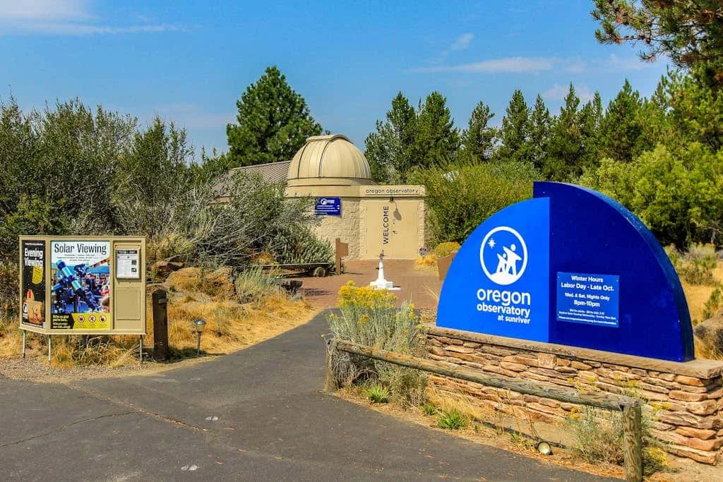 Entrada de un observatorio al aire libre, con un letrero azul junto a un camino pavimentado.