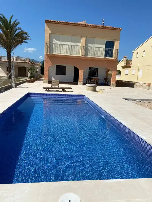 Una piscina frente a una casa de dos plantas bajo un cielo azul despejado.